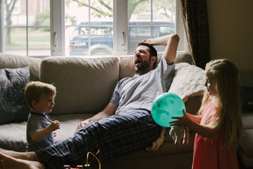 A tired father lies on the couch while his children play around him.