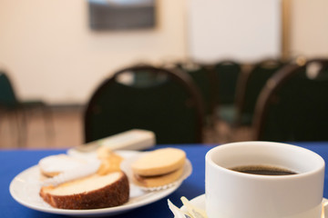 Break table with white cup of coffee and snacks over a blue table cover and conference room in the back