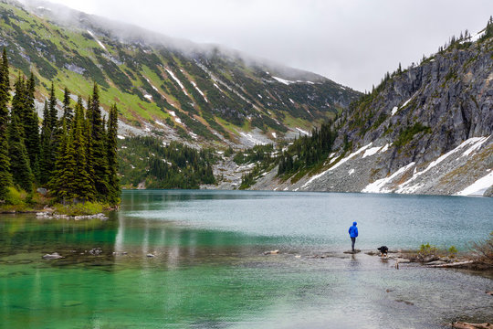 A women hangs out on the side of an alpine lake with her dog and takes in the view in the Coast Mountains around Pemberton, British Columbia, on a rainy summer day.