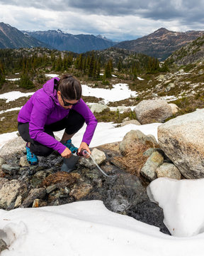 A women crouches down and uses a water filter to get drinking water from a small alpine stream in the mountains of British Columbia.