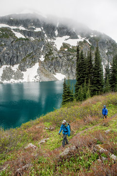 A man and women hike around a beautiful alpine lake on a cloudy, rainy summer day in the Coast Mountains around Pemberton, British Columbia.