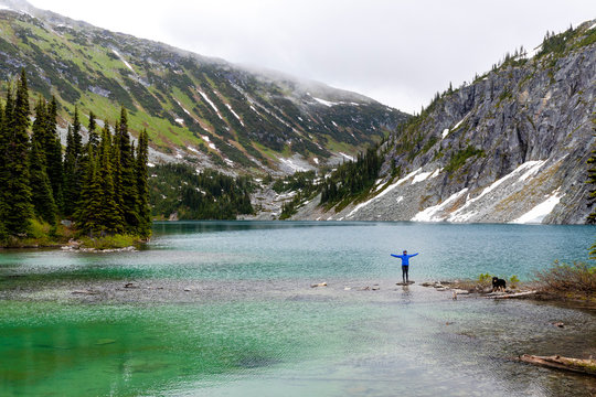 A women hangs out on the side of an alpine lake with her dog and takes in the view in the Coast Mountains around Pemberton, British Columbia, on a rainy summer day.