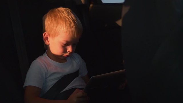 Little boy in the back seat of a car with a tablet in his hands. boy wearing a seat belt. Car travel with a tablet in hand