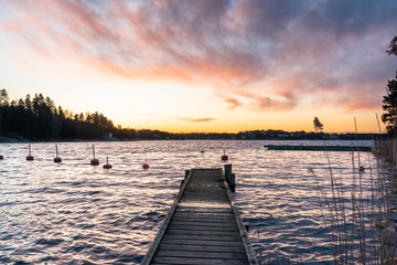 Fototapeta premium Old wooden jetty or pier background on the seaside. Sea bay at sunset. Horizon line, skyline with beautiful clouds in the rays of the setting sun. Colorful buoys floating on the surface of the water.