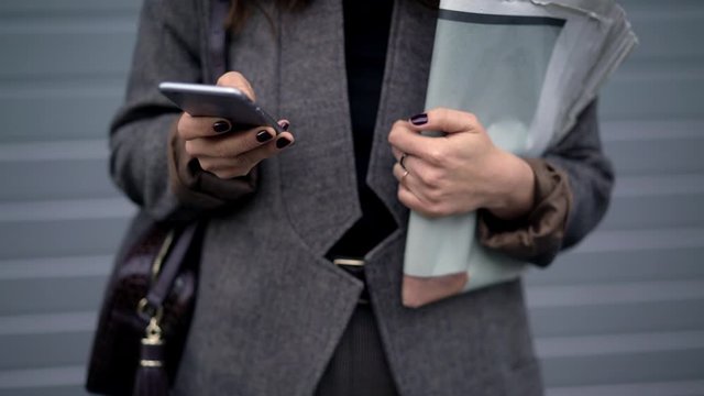 Pan shot left to right of woman wearing grey coat and bag, no face, holding newspapers and typing in the phone, standing on grey backgroud outdoors in city