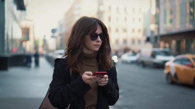 Slow motion of woman in sunglasses with a red phone on background of city road and car passing. Gimbal shot of woman typing in the phone walking along the road