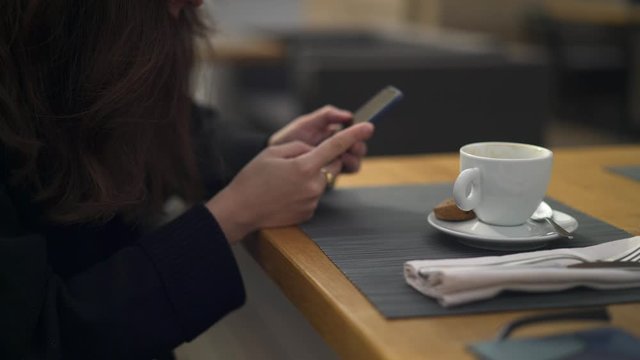 Pan shot left to right of woman no face holding typing on the phone drinking a cup of coffee at the cafe outside. Woman with a phone sitting alone at the restaurant drinking a cappuccino