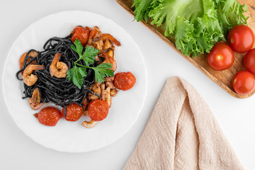 White plate with black spaghetti Nero and seafood, green lettuce leaves and cherry tomatoes on a white background top view
