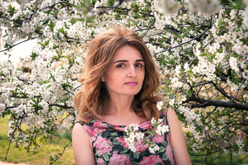 soft focus of attractive woman with long hair in dress with floral print looking at camera near blooming cherry tree with white flowers in garden