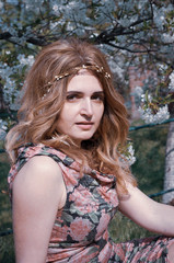 soft focus of attractive woman with long hair in floral wreath looking at camera in front of cherry blossoming tree in garden