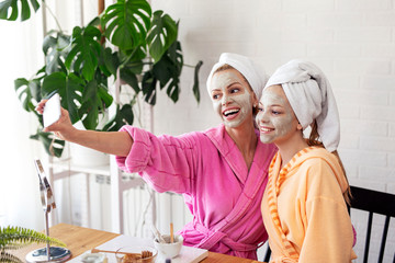 Mother and her daughter taking selfie by phone with facial mask and towel on head