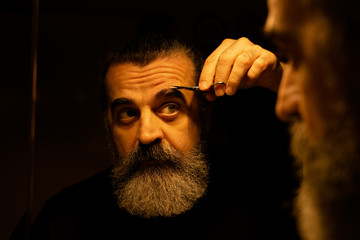 Bearded man grooming eyebrows, plucking hair with tweezers in front of mirror in dark room
