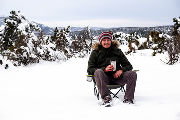 Handsome young man is sitting on camping chair, drinking coffee with thermos mug and enjoying snow in winter season. Winter in Eskisehir. - Powered by Adobe
