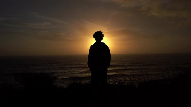 This stock video shows a silhouette of a man watching the sunset in a cliff over the sea.