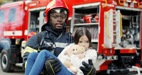 Portrait of african american brave fireman holds saved girl in his arms standing near fire truck. Firefighter in fire fighting operation