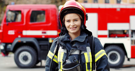 Portrait of young woman firefighter standing near fire truck. Fireman in protective suit with...