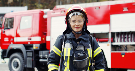 Portrait of young woman firefighter standing near fire truck. Fireman in protective suit with...
