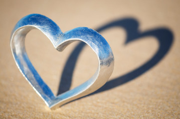 Shiny silver heart standing on the beach casting a shadow on brown sand