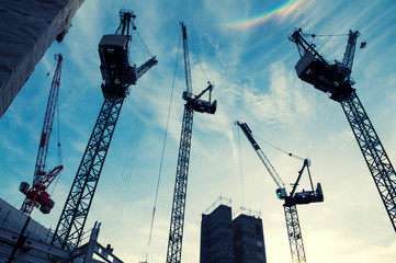 Construction cranes soaring in silhouette against blue sky with a rainbow flare in the clouds 