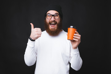 Young man with beard is showing exited a thumb up and holding a cup with hoe water.
