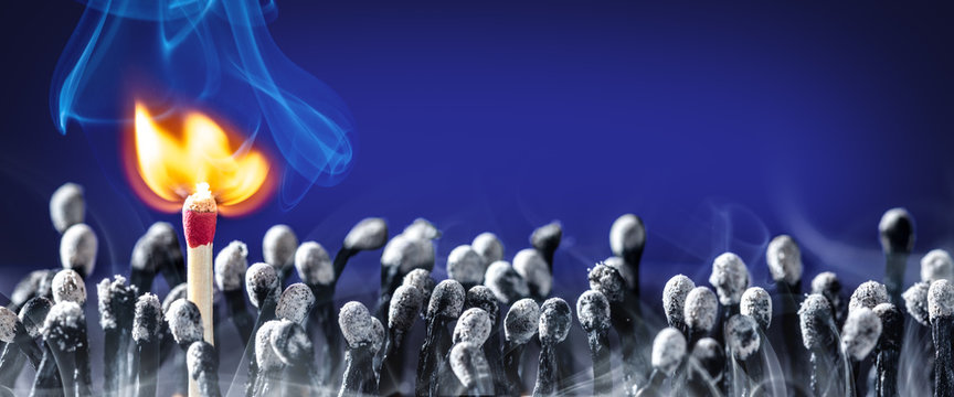 New Match Bursting Into Flames Standing In Group Of Old Burnt Matchsticks - New Life / Idea Concept