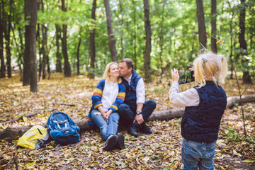 Fototapeta premium travel, tourism, hike, technology and family concept. Happy child makes photo parents in forest. Mom and dad pose for photo, daughter takes photo on phone. Young family of tourists in wooded area