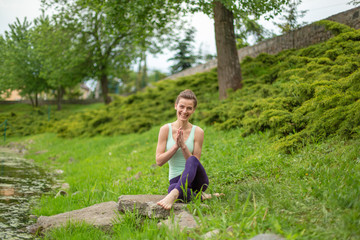 Naklejka na ściany i meble A young sports girl practices yoga on a green lawn by the river, yoga assans posture. Meditation and unity with nature