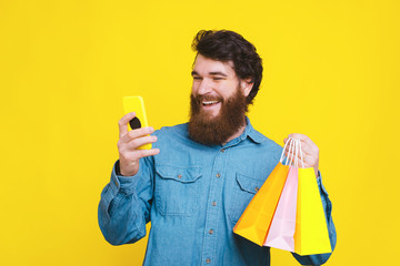 Photo of happy bearded hipster man using smartphone and holding shopping bags