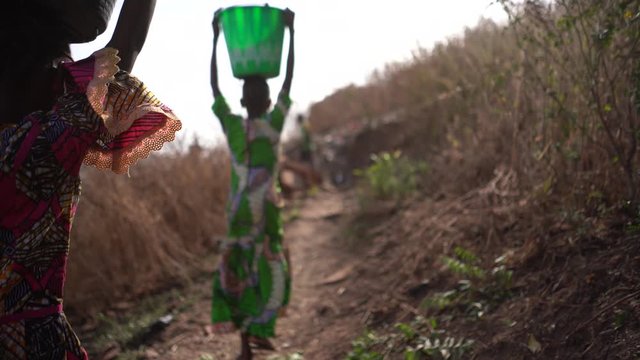 Back View Of Two African Girls With Water Buckets On Their Head Walking Climbing Up A  Mountain Trail