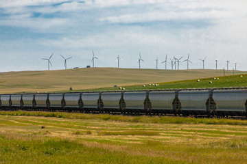 Row of metallic wagons of a Canadian freight train running between green fields in the countryside. Wind turbines in the background.