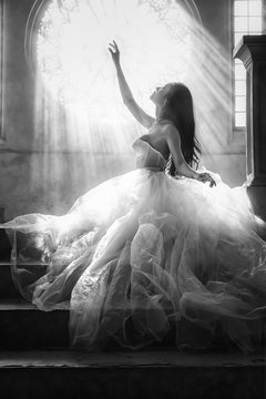 silhouette black and white portrait of bride in white dress sitting on stairs infrontof window with sunlight pass through