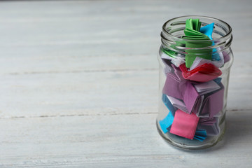 Glass jar with colorful paper pieces on white wooden table, space for text