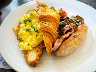 All-Day breakfast / brunch meal platter of scrambled eggs; croissant; sausage; bacon and mushrooms on a white plate