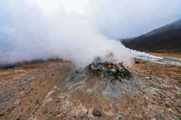 Hot sulfuric steam vent spewing sulphur steam in the hot sulfuric and geothermal area of Namaskard in Myvatn/Iceland. Color and mineral rich textured muddy ground infront.