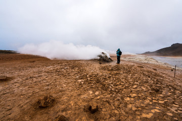 Fototapeta na wymiar Girl with hiking gear infront of sulphur steam vents in Iceland during heavy cold wind. Mineral rich and textured muddy ground infront. Geology, traveling and volcanic energy concept.