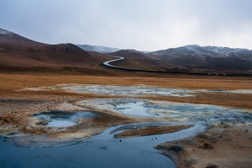 Textured, cracked and patterned ground in the Namaskard/Myvatn area in Icelandic nature. S shaped road and snow covered mountains in the background. Minerals and volcanic energy concept.