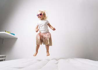 A little girl in pink sunglasses and a pink skirt jumps on a white mattress.