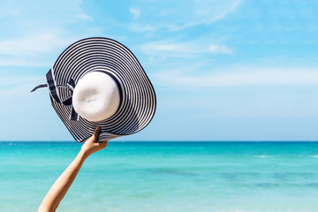 Summertime.  Close up raise hand travel women holding big fashion hat on the sand beach for summer and vacations holiday, blue sky and beach background. Summer and Travel Concept