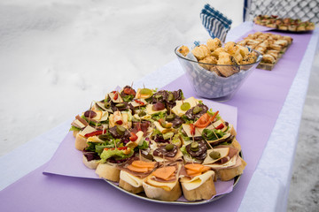 Buffet with sandwiches made of bread, cheese, salami, ham, prosciutto, olives,.. in winter setting on purple table on a ski slope.