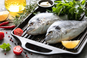 Two raw dorado fish with spices, lemon and parsley in a black grill pan on a white concrete background.