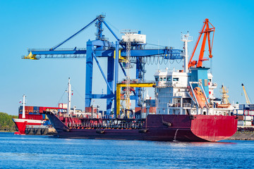 The ship is waiting for loading at the sea commercial port. Ship on the background of cranes. The red ship is moored. Shipping. Freight. Cargo transportation by sea.