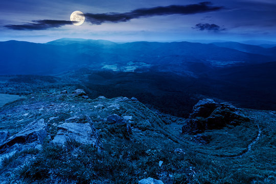 mountain landscape in summer at night. view from the top of carpathian watershed ridge in to the distance in full moon light. boulders on the grassy slopes. weather with clouds on the dark sky