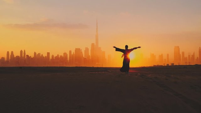 Arabic woman weared in traditional UAE dress - abayain rising her hands on the sunset at a desert with Dubai city silhouette on the background.