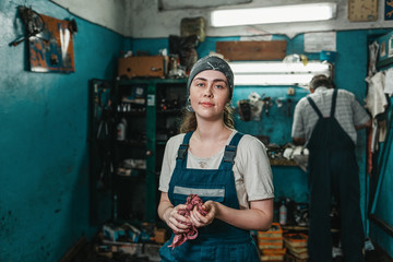 Fototapeta na wymiar Gender equality. Portrait of a young smiling woman in uniform working in a workshop, who wipes her hands with a rag. In the background, a worker at the machine
