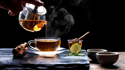 Pouring hot aromatic herbal tea from teapot into glass teacup set with steam and various herbs on...