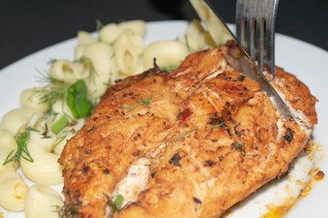 Chicken breast fried with spices, boiled pasta with herbs, metal fork and knife which cut a piece of the breast on a white plate.