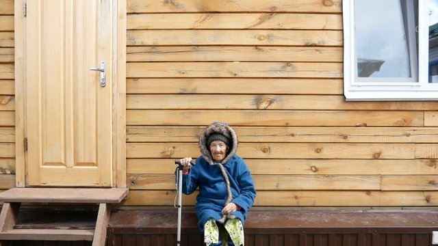 Grandma is holding a wand and sitting next to the house, looking straight ahead, she is wearing warm clothes, sitting on a green background.