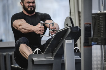 strong young man with beard in black sportswear pulling heavy weight sitting in exercise equipment...