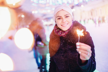 Happy young woman with winter clothes smiles lights sparklers, background evening city illumination. Christmas concept