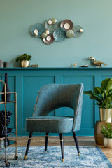 Interior design of living room with stylish  armchair, gold liquor cabinet, decoration, pillow plants and elegant personal accessories. Design home decor. Green wall panelling with shelf. Template. 